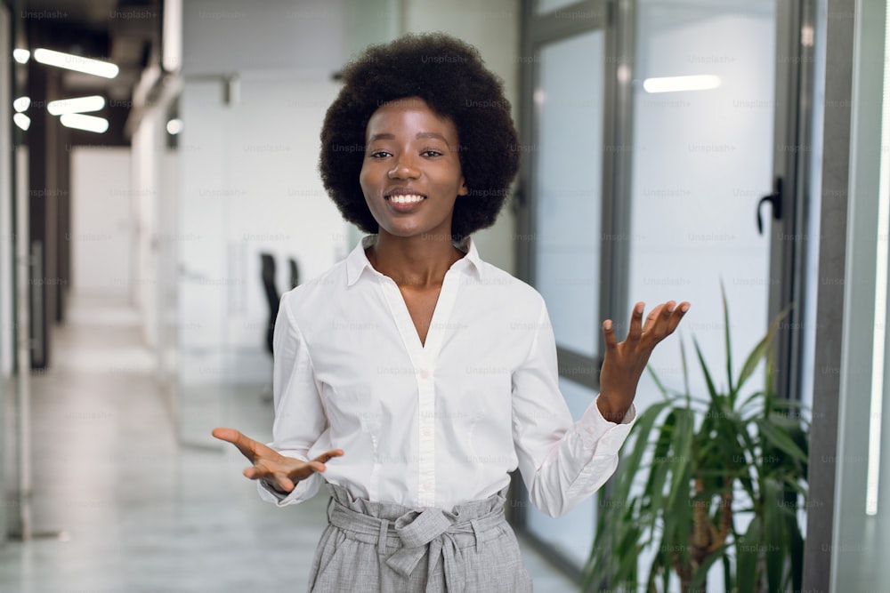 Joven atractiva y exitosa mujer de negocios afroamericana negra en ropa formal, dando una entrevista, hablando y gesticulando, mientras está de pie en el interior de un moderno edificio de oficinas.