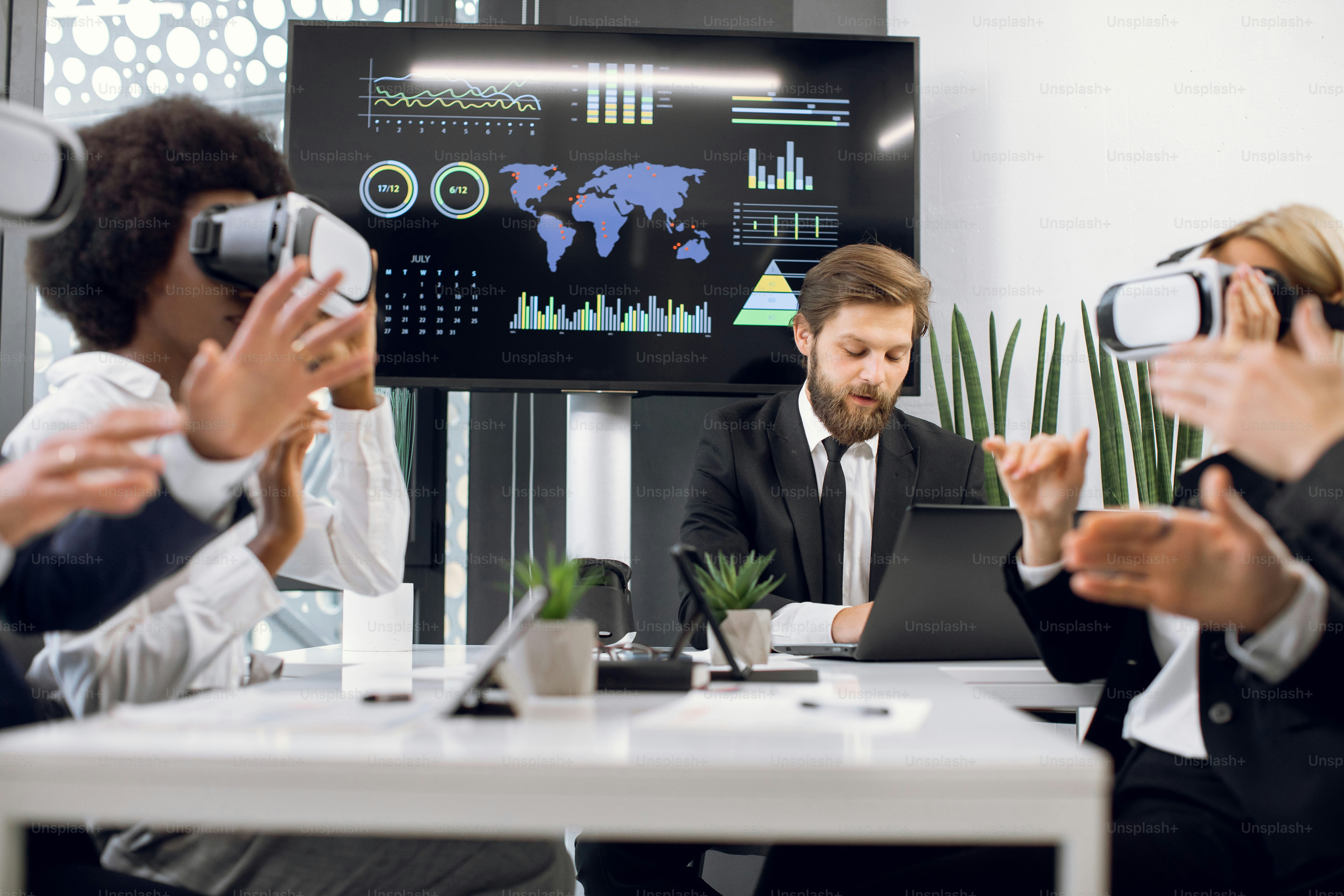 Handsome male bearded businessman, sitting in front of plasma screen, presenting new project to her creative multiracial team, using virtual reality simulators and gesturing.