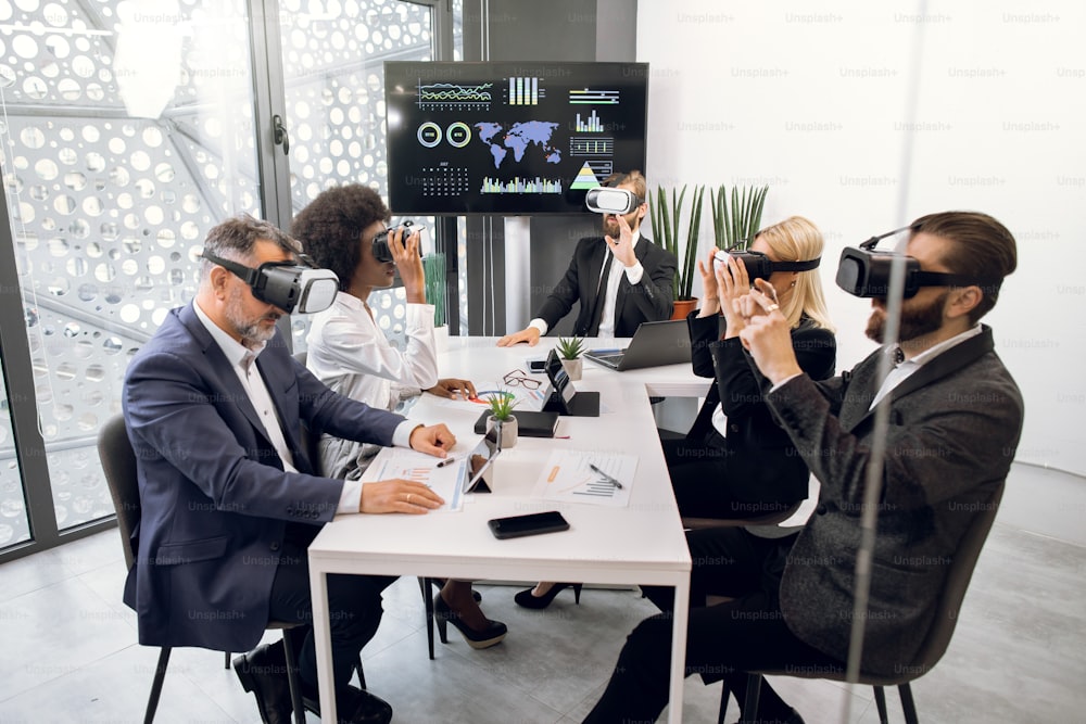 Focused five multiethnic professional business colleagues in formal wear, sitting at the table in office and using VR headsets. Business, technology, testing of mobile applications concept.