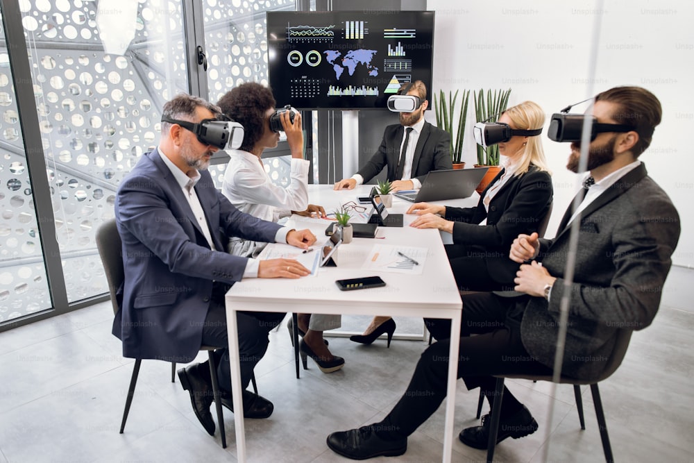 Team aziendale creativo multietnico che utilizza cuffie per realtà virtuale durante la riunione in un ufficio moderno. Gruppo di sviluppatori che utilizzano simulatori di realtà virtuale, seduti intorno al tavolo in ufficio