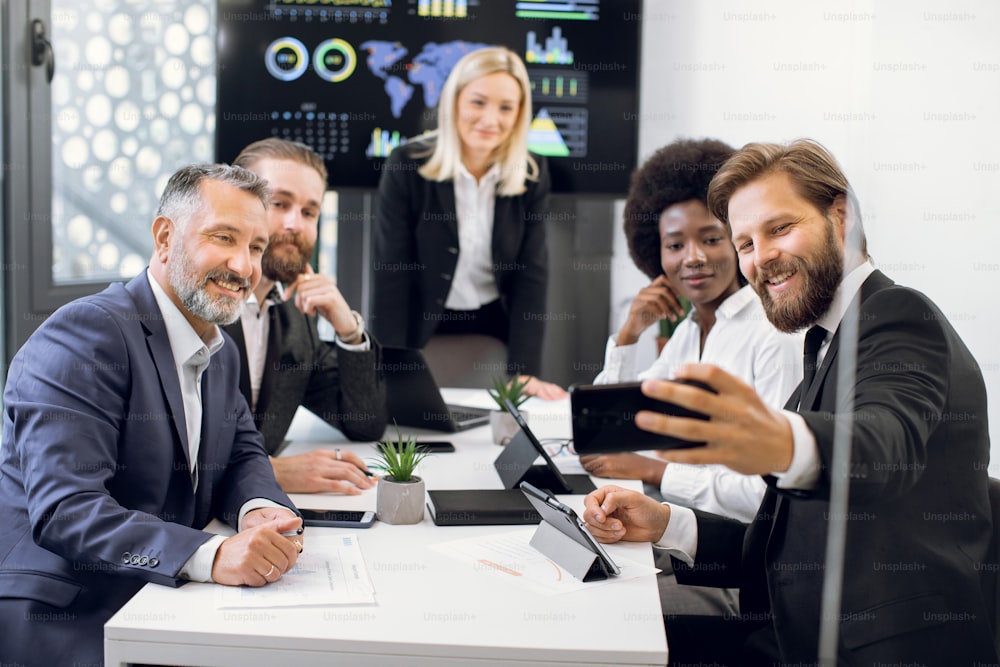 Team of good-looking smiling successful diverse corporate coworkers with mature female boss, posing on phone camera for selfie photo, during meeting in office room with big wall screen.