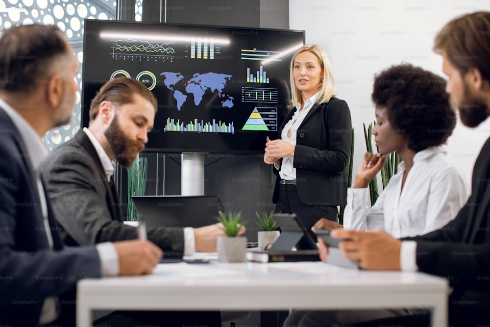 Middle aged pleasant confident blond business lady, showing company's work infographics on digital wall screen during meeting with team of diverse multiracial male and female businesspeople.