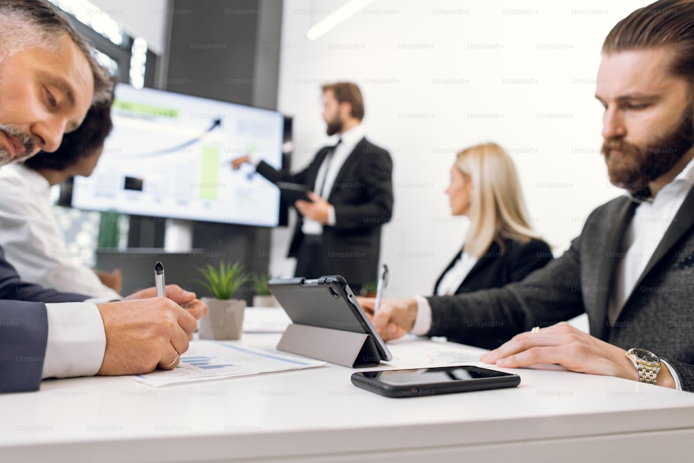 Due uomini d'affari diversi focalizzati, che prendono appunti mentre ascoltano il discorso dell'oratore maschio, mostrando la presentazione finanziaria a diversi gruppi di dipendenti alla riunione dell'ufficio. Concentrati su tablet e telefono.