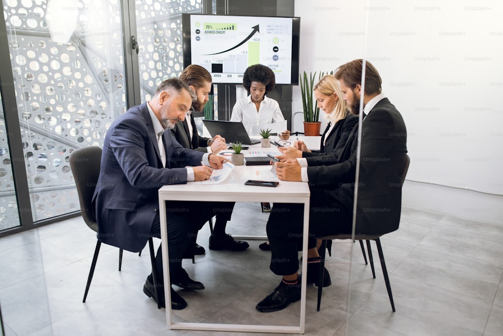 Group of concentrated high-skilled confident multiethnic business colleagues, sitting at the table in conference room and working together over business strategy of company.