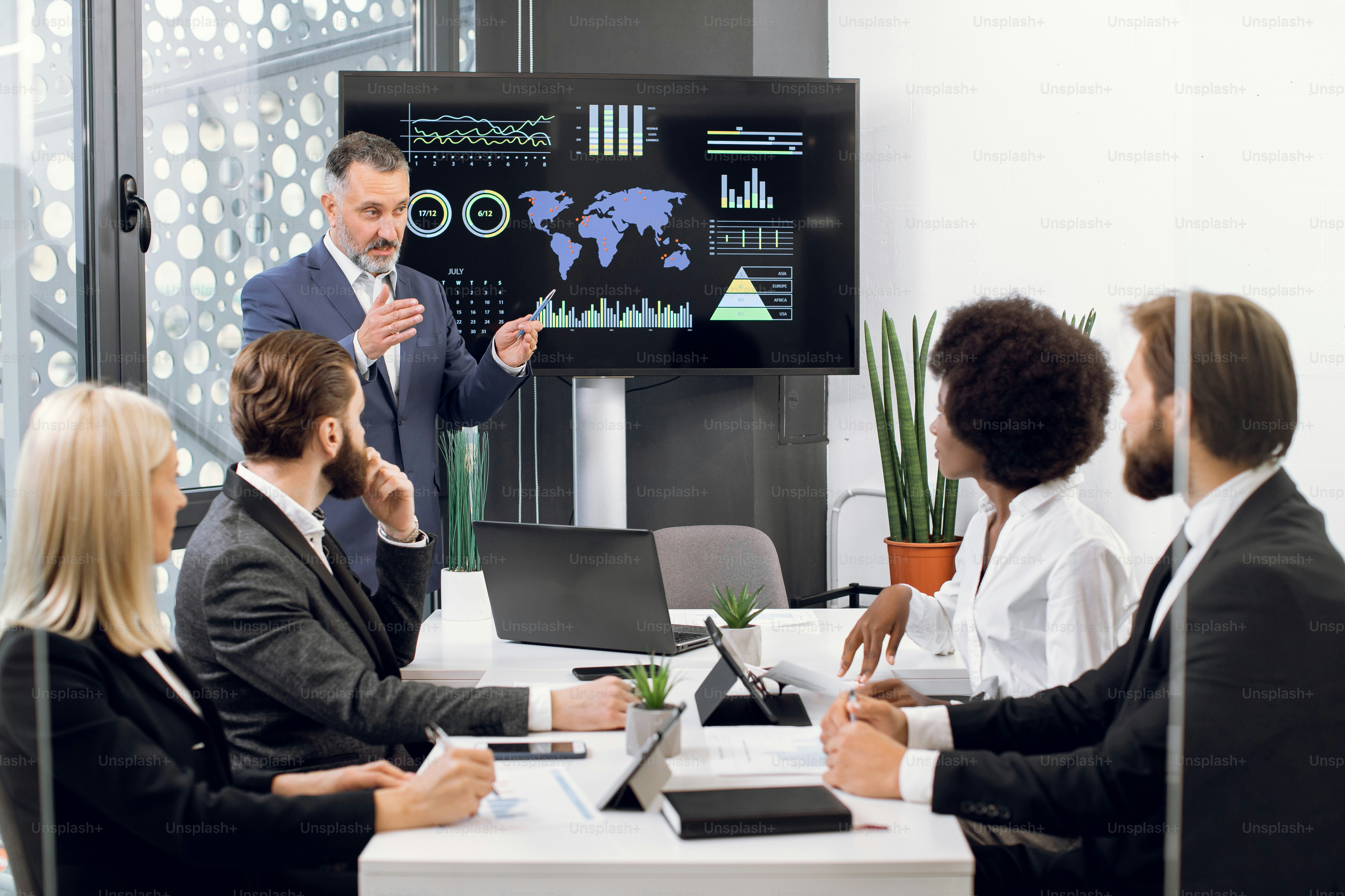 Serious mature businessman manager stands near big digital wall screen with company financial and growth charts and talks to his multiethnic colleagues, discussing results of the project.