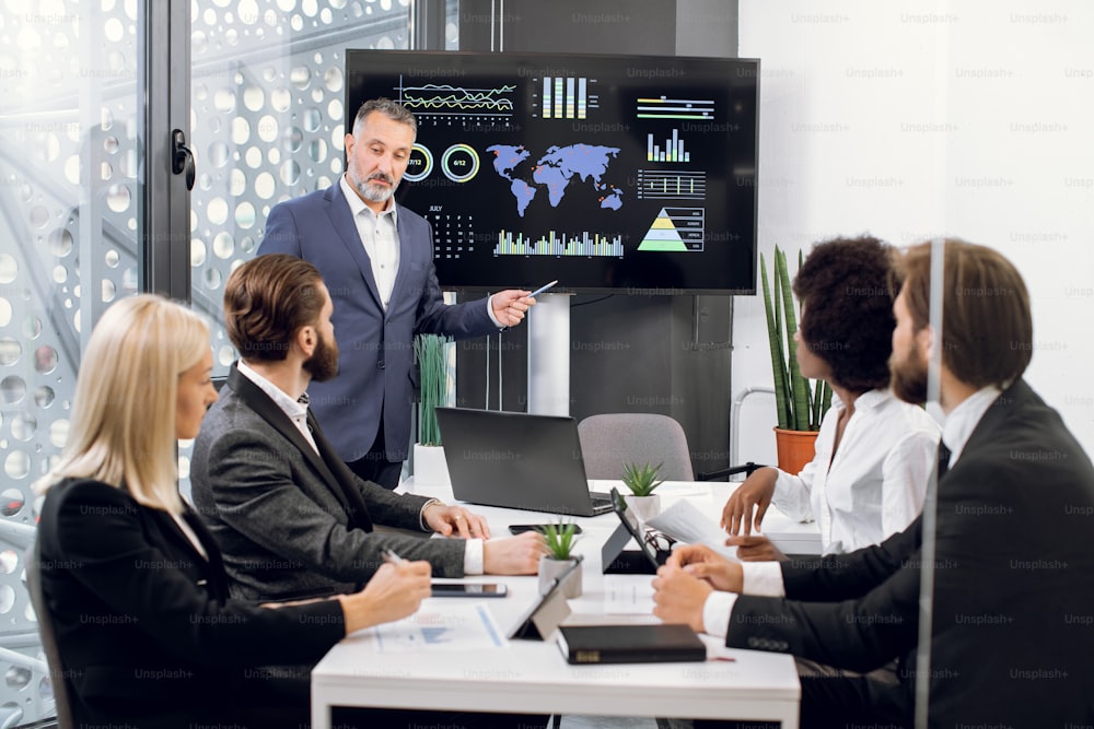 Male mature manager in business suit, speaking at office meeting with his multiethnic male and female colleagues, sitting at table, showing company's annual growth report charts and map on TV screen.