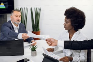 Cropped close up shot of young African businesslady, sitting at the table in modern light office between two male colleagues, giving paper report each other. Business meeting, brainstorming.