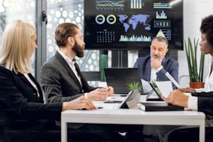 Team of multiracial concentrated businesspeople having meeting in boardroom at office, sitting at the table in front of a huge plasma TV screen. Focus on bearded man talking to colleagues.