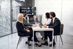 Équipe de cinq hommes d’affaires multiethniques se réunissant dans une salle de conférence au bureau à l’intérieur, assis au bureau avec des tablettes et un ordinateur portable, devant un immense écran de télévision à plasma.