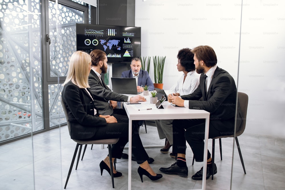 Team of diverse five multiethnic businesspeople having meeting in boardroom at office indoor, sitting at the desk with tablets and laptop, in front of a huge plasma TV screen.