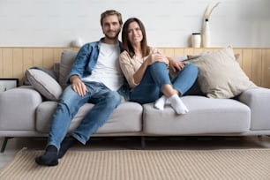 Happy young Caucasian couple relax on cozy sofa at home hugging look in distance dreaming together, dreamy millennial man and woman rest on comfortable couch embracing, enjoy weekend at home