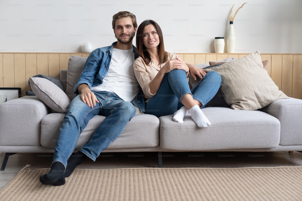Happy young Caucasian couple relax on cozy sofa at home hugging look in distance dreaming together, dreamy millennial man and woman rest on comfortable couch embracing, enjoy weekend at home
