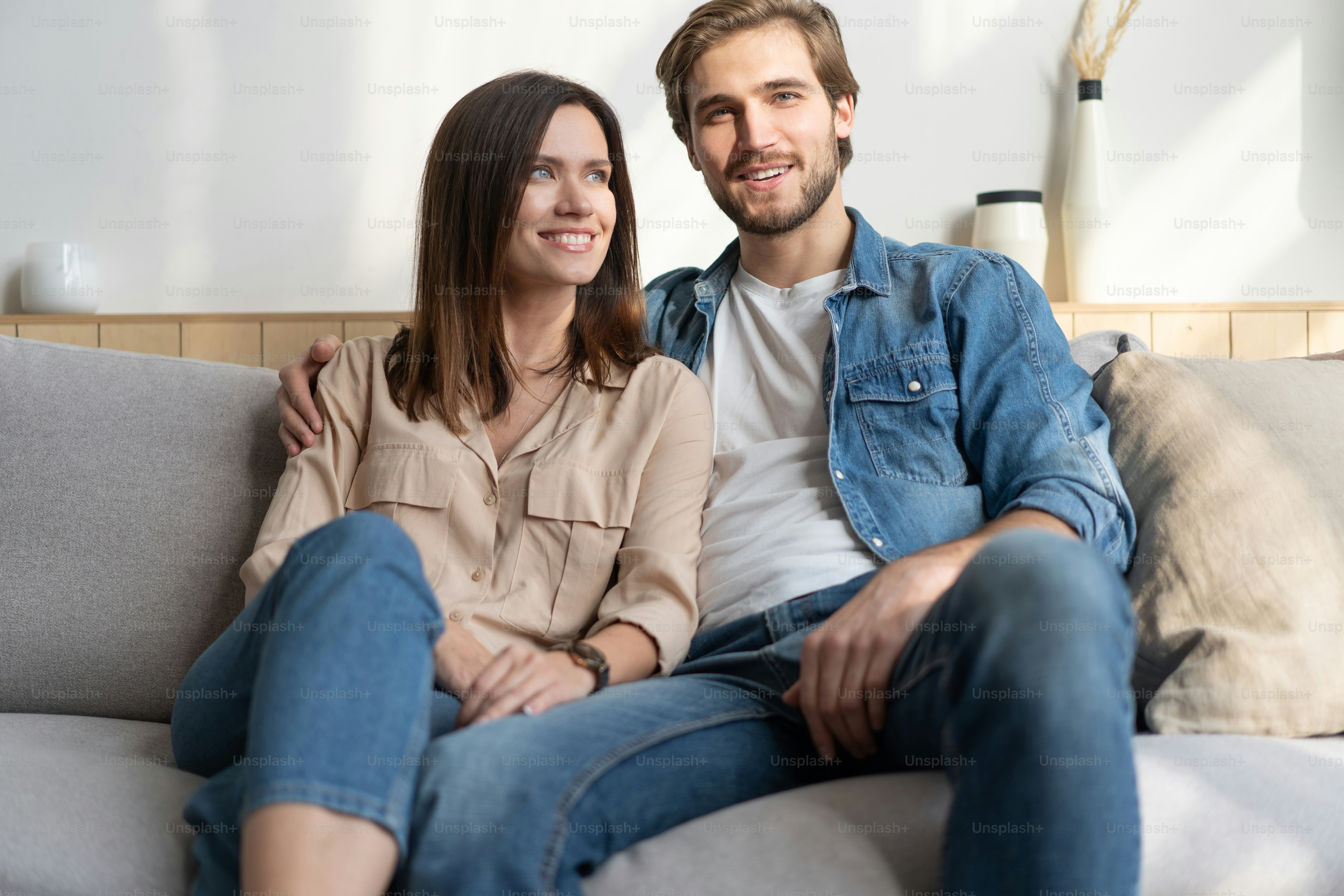 Happy young Caucasian couple relax on cozy sofa at home hugging look in distance dreaming together, dreamy millennial man and woman rest on comfortable couch embracing, enjoy weekend at home