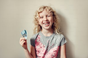 Caucasian blonde girl showing her missing tooth in mouth and holding money from tooth fairy. Proud child kid showing lost tooth. Growing up stage and adolescence.