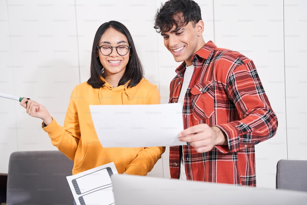 Fröhliche junge qualifizierte Designer stehen im Büro und diskutieren lächelnd über ihr Projekt. Mann hält Skizze des zukünftigen Projekts, während charmante Frau es mit positivem Lächeln betrachtet