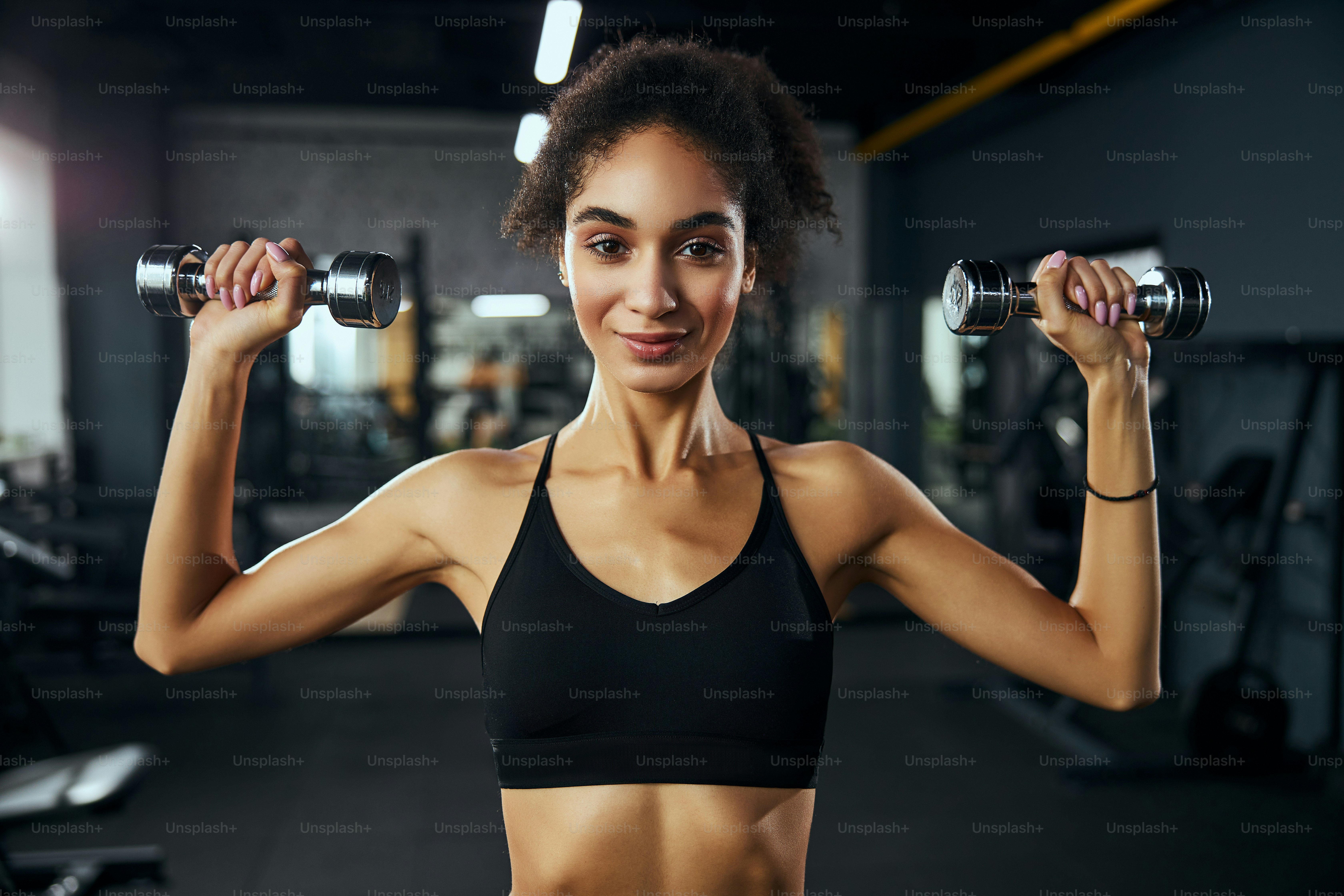 Attractive international female keeping smile on her face while lifting dumbbells