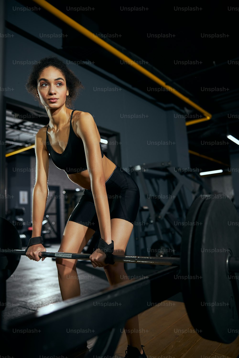 Kind international woman looking aside while lifting heavy bar, working at quality of her muscles