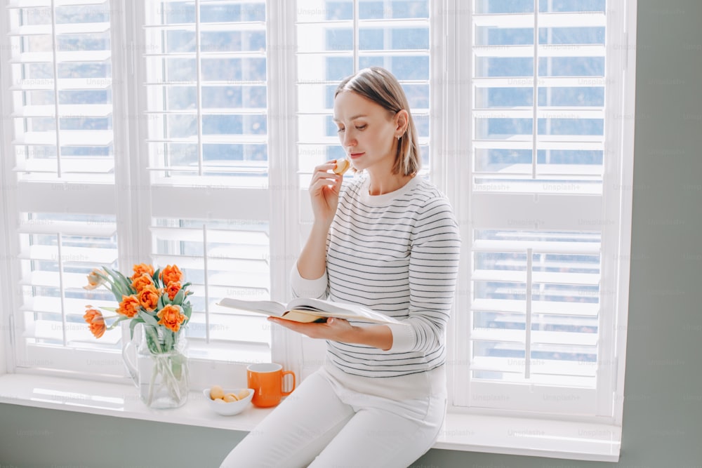 Beautiful happy middle age woman sitting on windowsill at home and reading book. Young woman with short hair eating macaroons and holding reading book. Relax, leisure time at home indoor.
