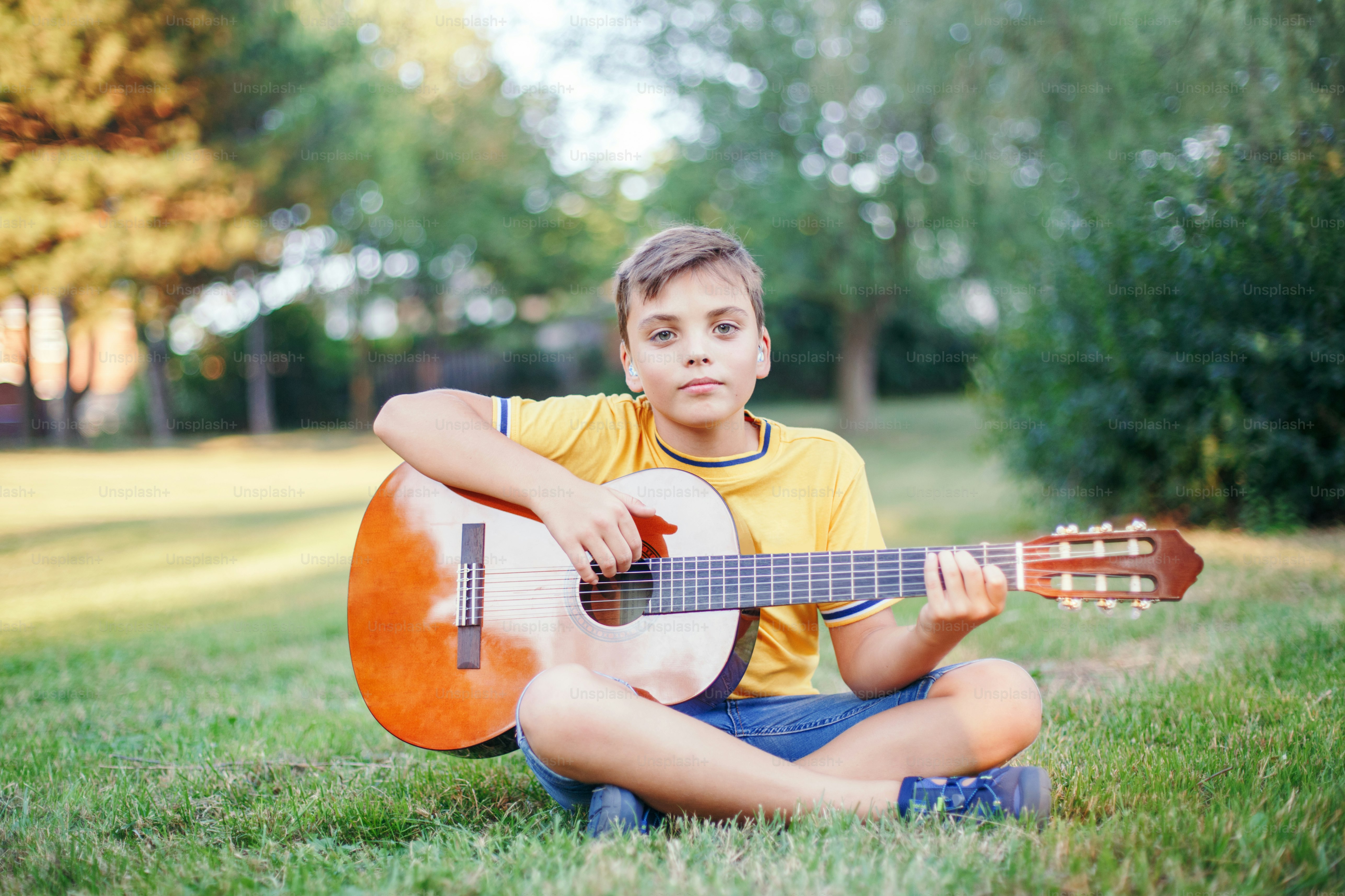 Niño preadolescente con problemas de audición tocando la guitarra al aire libre. Niño con audífonos en los oídos tocando música y cantando canciones en el parque. Actividad artística de pasatiempo para niños. Auténtico momento de la infancia.