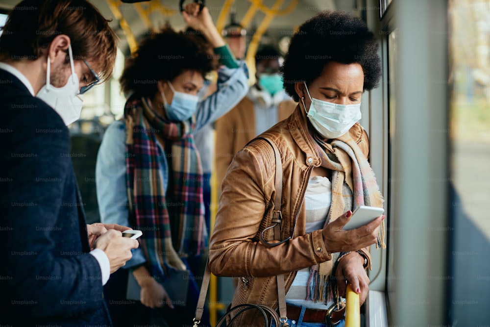African American woman using mobile phone while traveling by public transport and wearing face mask due to coronavirus pandemic.