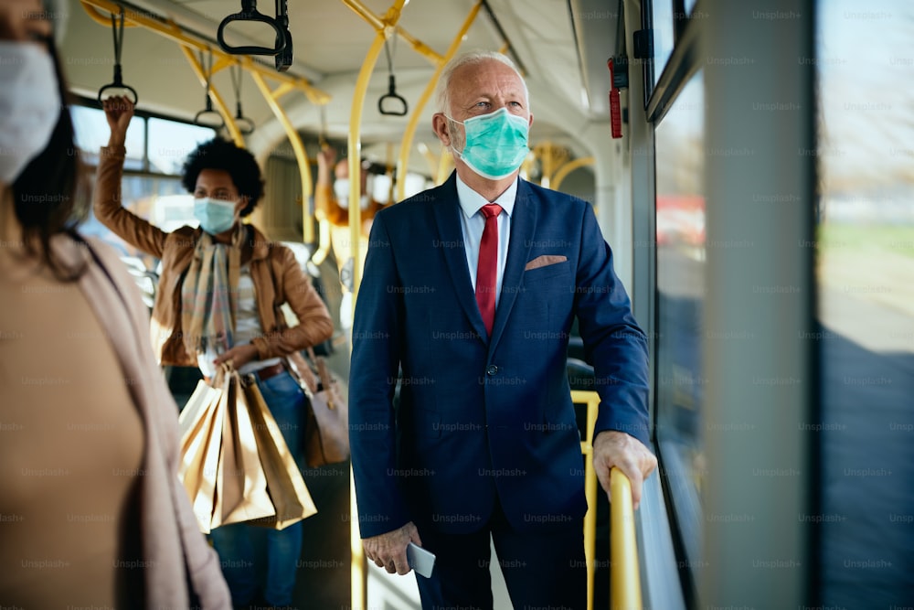 Uomo d'affari maturo con maschera facciale che si sposta in autobus e guarda attraverso il finestrino.