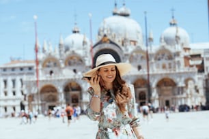 이탈리아 베니스의 산 마르코 광장에서 선글라스와 모자를 쓴 꽃무늬 드레스를 입은 행복한 현대 여행자 여성이 스마트폰으로 이야기하고 있다.