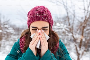 Mujer joven enferma y estornuda en papel de seda. Chica sonándose la nariz al aire libre. Resfriarse en invierno. Mujer triste molesta con mala expresión estornuda y tiene secreción nasal
