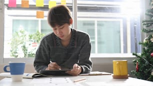 Young female creative designer working on digital tablet at her workspace near window with sun flare.