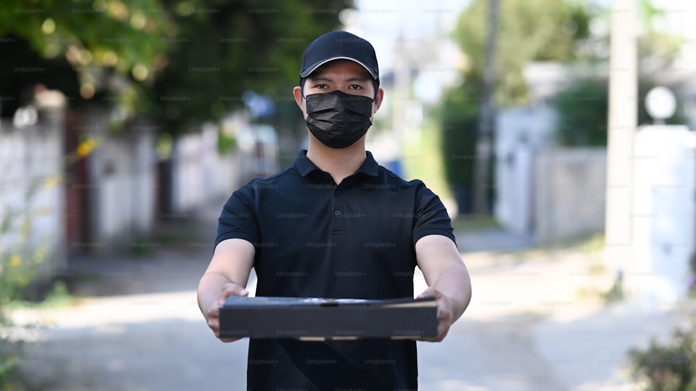 保護マスクを着けた若いアジアの配達員が、熱々のピザの入った箱を持っている。