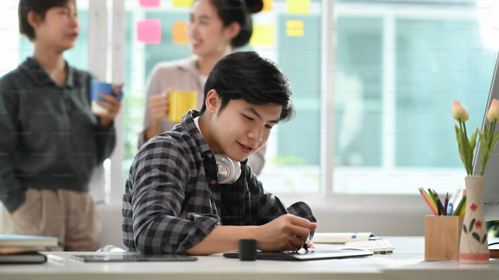 Young asian entrepreneur of small company man working on digital tablet at modern office with his colleagues standing in background.