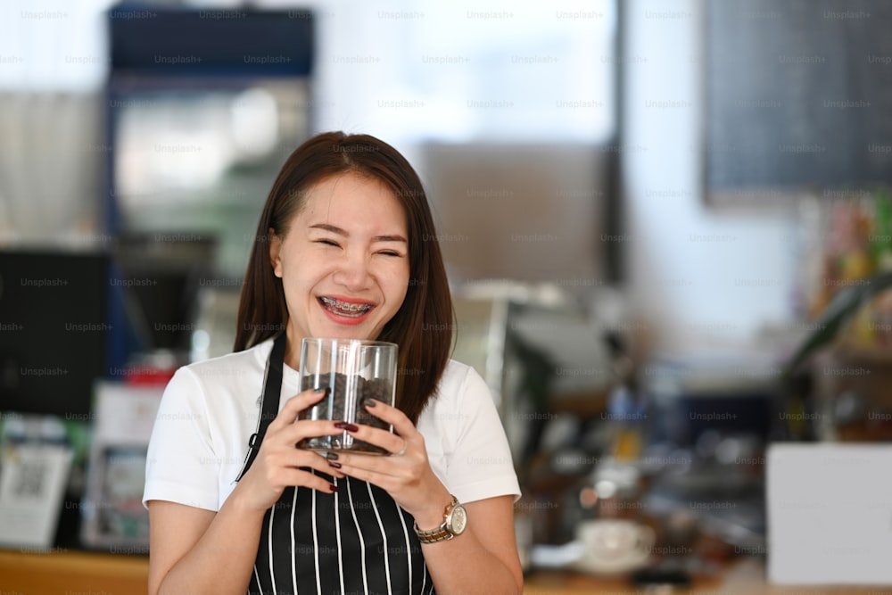 Happy Asian barista woman  holding glass bowl with fresh roasted coffee beans in coffee shop.