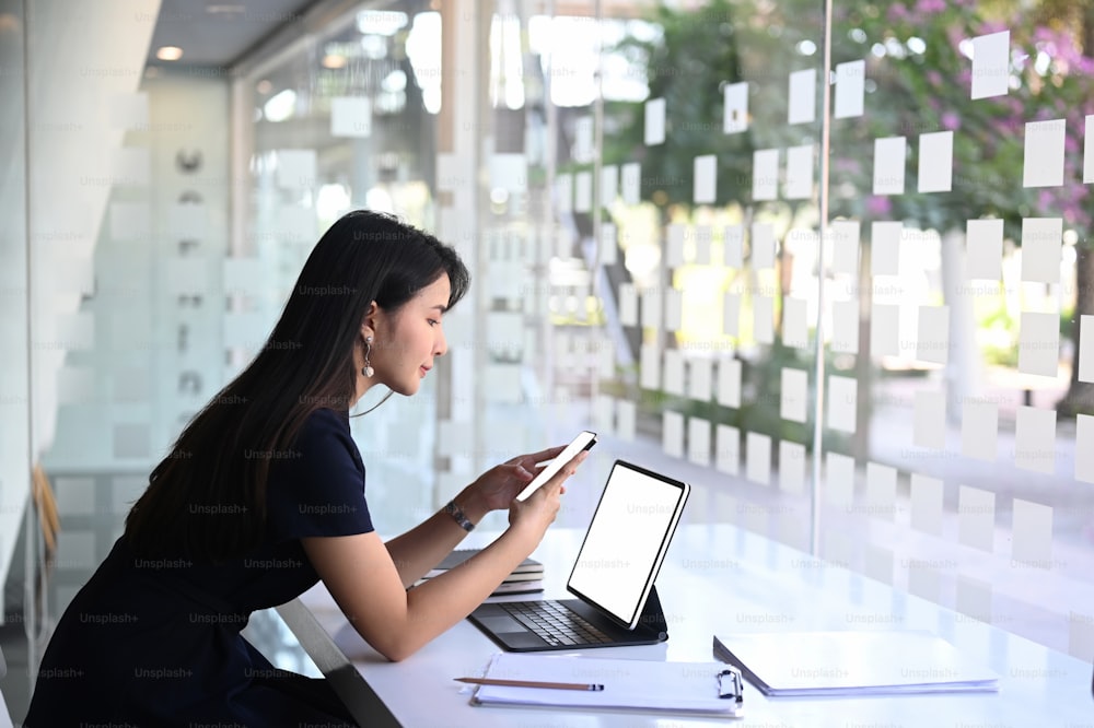 Side view of businesswoman searching information on smartphone and working with tablet computer at her workstation.