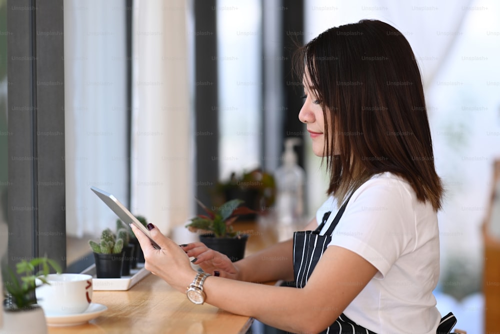 Mujer emprendedora con un delantal usando una tableta digital mientras está sentada cerca de la ventana de su cafetería.