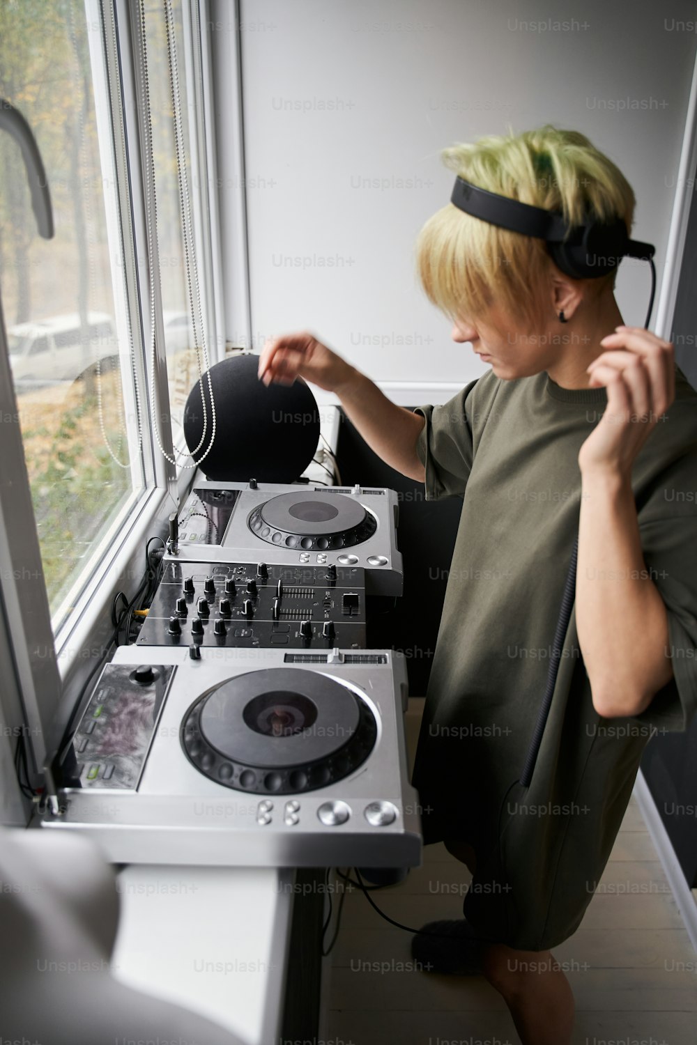 금발의 힙스터 여자는 홈 스튜디오에서 음악을 녹음하고 믹싱합니다. 아늑한 방에서 창턱에 서서 음악 트랙을 만드는 어린 소녀