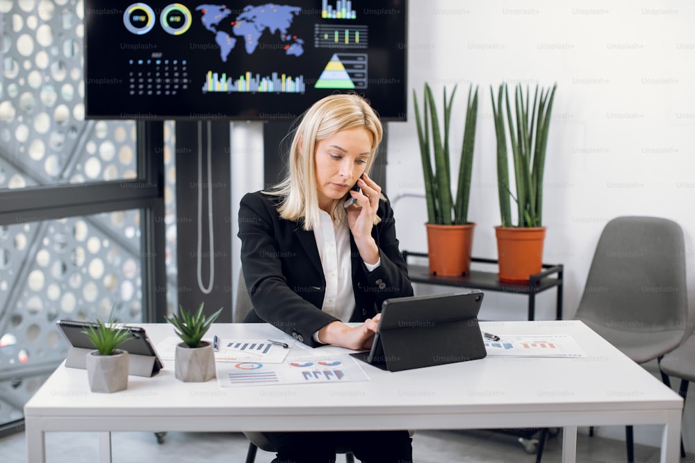 Horizontal shot of attractive focused high-skilled mature blond businesswoman which talking on phone and simultaneously typing on digital tablet, sitting at the desk in board office room.
