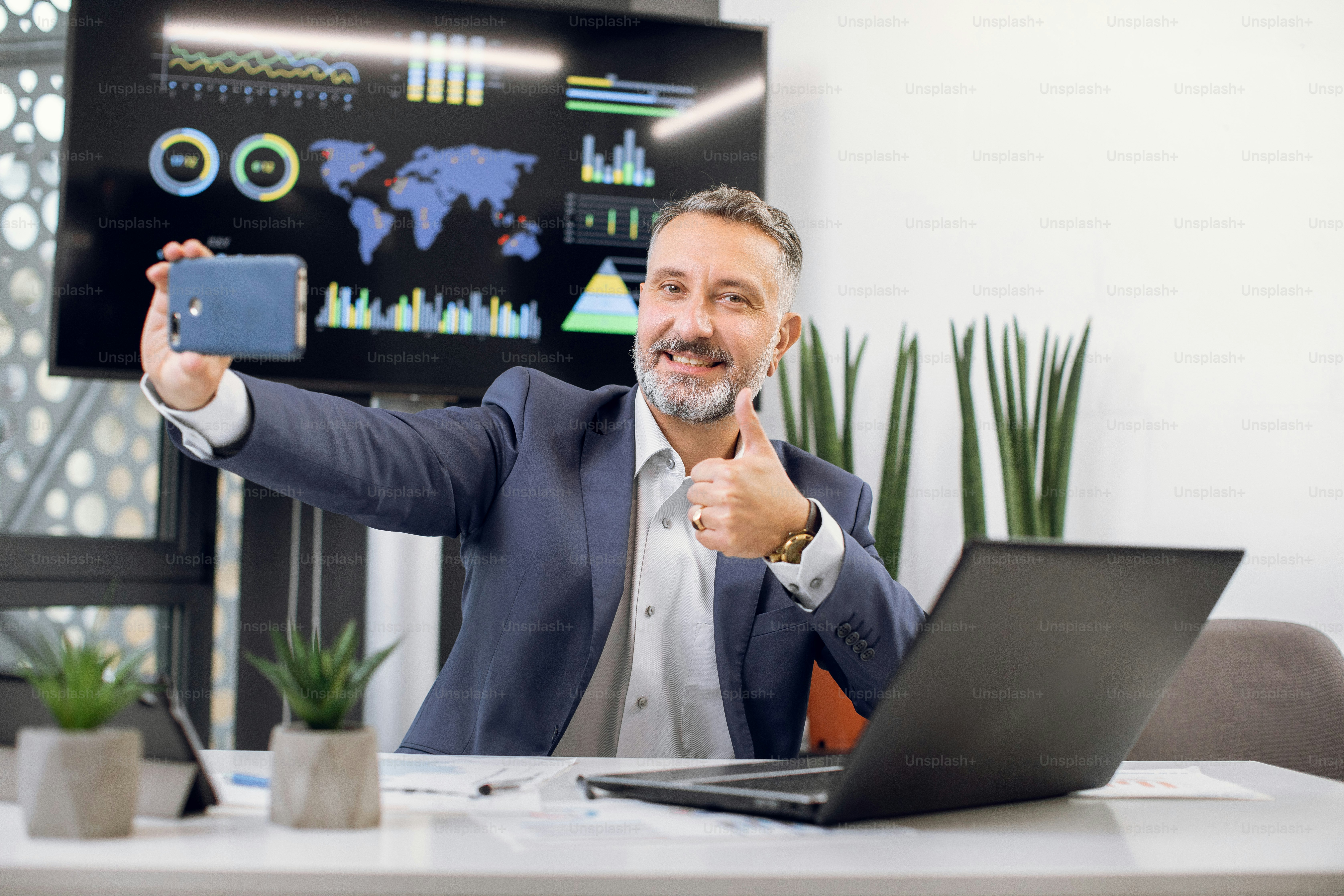 Close up portrait of pleasant mature bearded businessman with gray hair, making selfie photo and showing thumb up, while working on laptop in modern office interior.