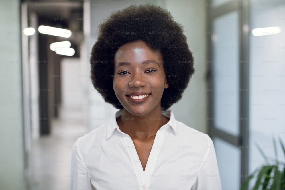 Close up face portrait of pretty smiling black young woman with afro hair, wearing white shirt, looking at camera, while standing inside modern office building. Businesswoman indoor portrait.