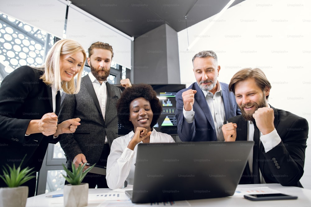 Successful team of multiethnic business people are raising hands in fists and screaming with happiness while working with a laptop computer in business center. Business people clenching fists.