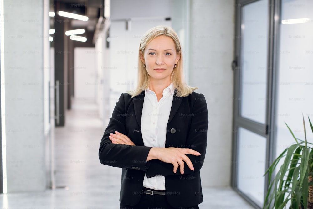 Indoor office portrait of pretty mature blond business lady, wearing white shirt and black jacket, standing with arms crossed in modern office interior. Businesswoman in office corridor.