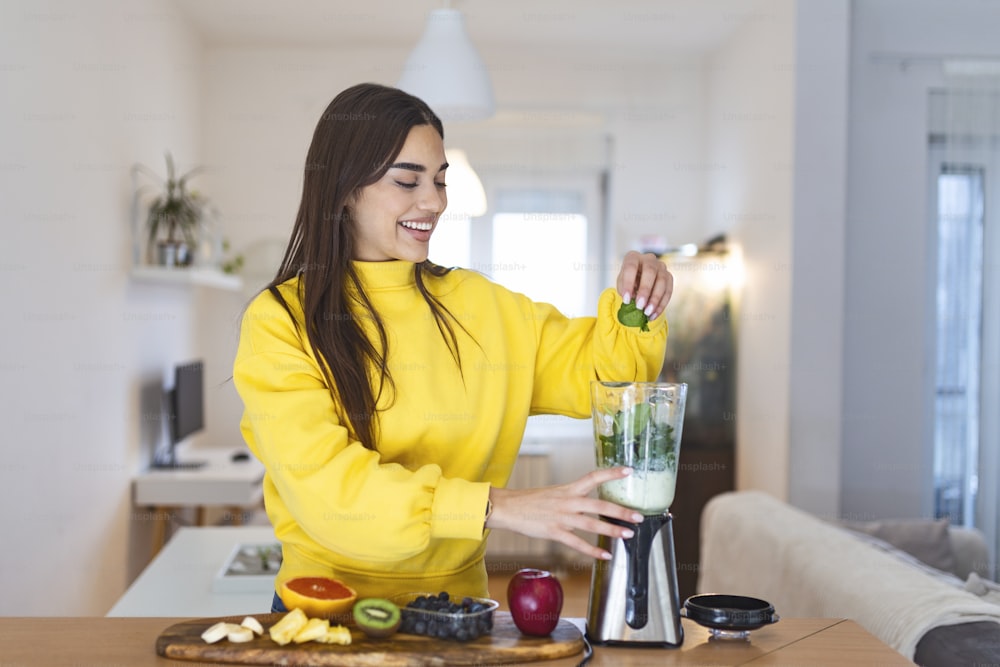 Mulher nova fazendo smoothie de desintoxicação em casa. Menina que faz smoothie de frutas e legumes. Modo de vida saudável. Menina feliz usando o liquidificador para fazer smoothie