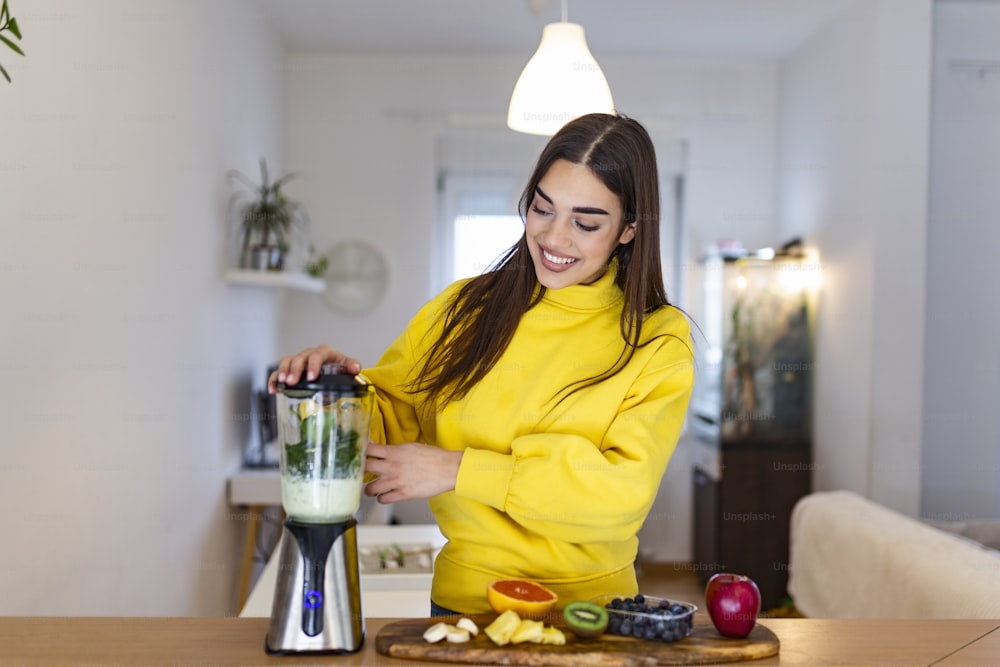 Mujer joven haciendo batido con frutas. Hermosa chica de pie en la cocina y preparando batido con frutas y verduras