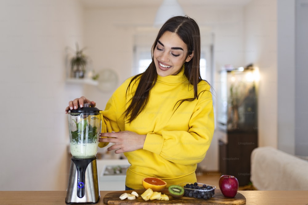 Young woman making smoothie with fruits. Beutiful girl standing in the kitchen and preparing smoothie with fruit and vegetables