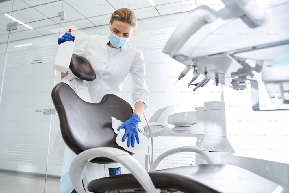 Low angle view of the female blonde dentist carefully sterilize the medical armchair inside a dental clinic during the coronavirus pandemic. Working during pandemic concept. Stock photo