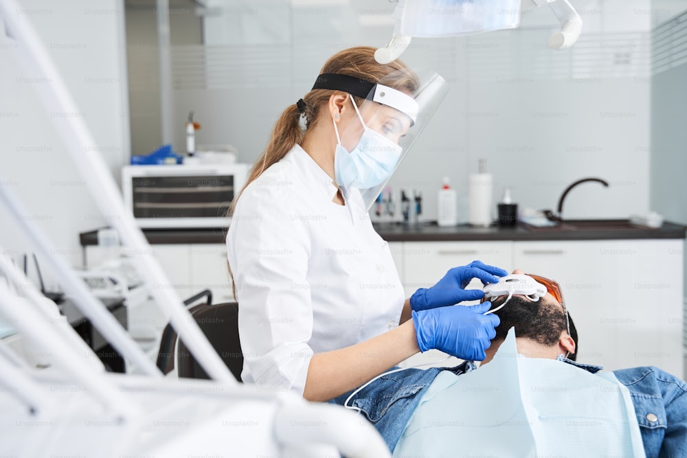 歯科用UVホワイトニング装置で歯を白くする男性、患者の世話をする歯科助手、眼鏡で保護された目。光、レーザー、フッ化物による美白治療。人工歯のホワイトニング