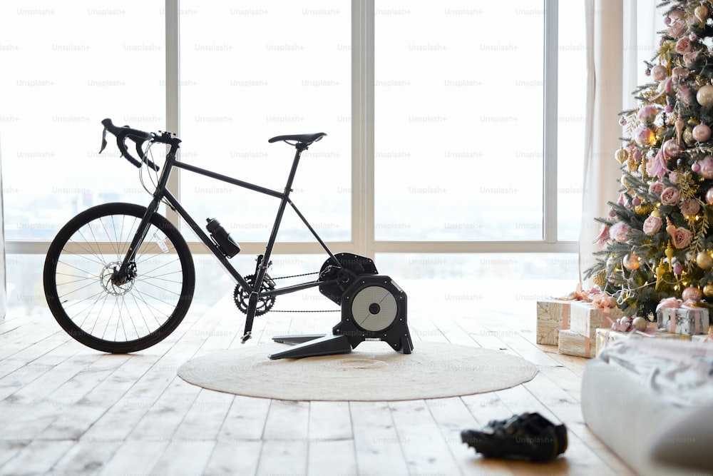 Vélo d’appartement noir ou vélo stationnaire dans la chambre avec de grandes fenêtres et des cadeaux de Noël sous le sapin