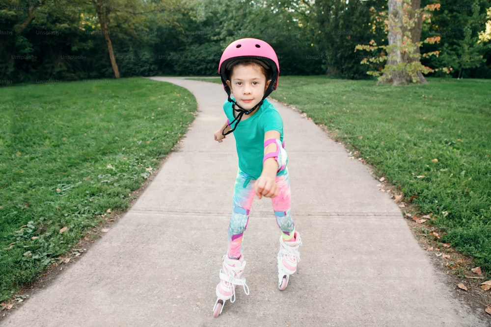 여름날 공원의 도로에서 롤러 스케이트를 타는 분홍색 헬멧을 쓴 행복한 백인 소녀. 계절에 따라 야외 어린이 활동 스포츠. 건강한 어린 시절의 생활 방식. 어린이 개인 여름 스포츠.
