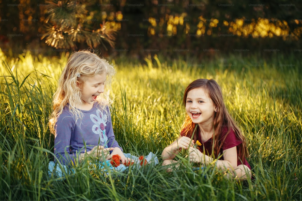 Happy children girls playing dolls in park. Cute adorable kids sitting in grass on meadow playing toys. Happy childhood authentic lifestyle. Outdoor summer backyard activity for kids.