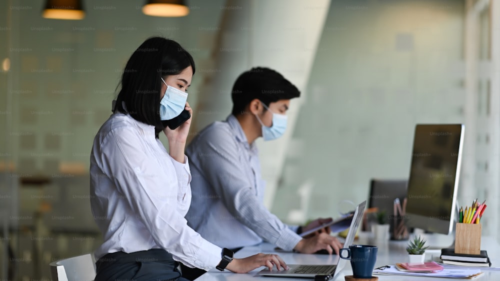 Side view of asian office employee woman wearing protective face mask work in new normal office with her colleague.