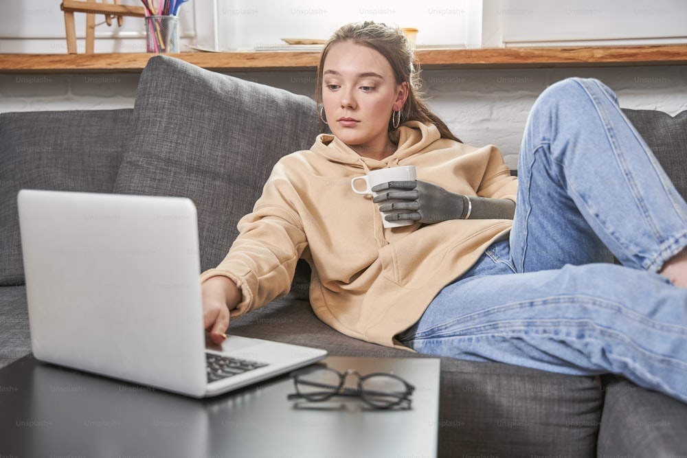 Portrait of an excited ginger woman with artificial limb relaxing on a couch at home while watching movie at the laptop and drinking beverage. Woman relaxing after home tasks. Disabled people concept