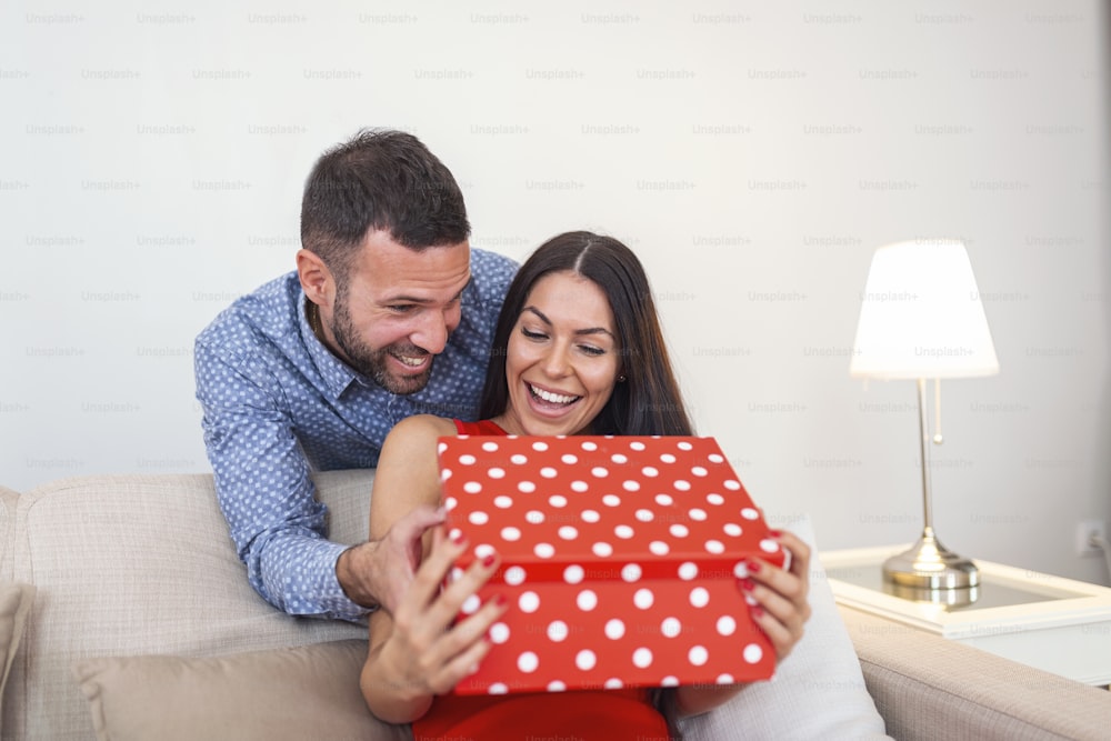Bofriend surprising his girlfriemd with a gift. Young man giving a gift box to his girlfriend. Valentine's Day and people concept - close up of young couple with gift box at home